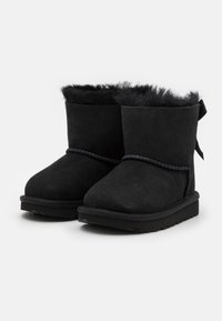 Bottines noires en cuir suédé doux avec un revers en fourrure bouclée, dotées d'un laçage décoratif et d'un bout arrondi. La semelle est texturée pour une meilleure adhérence.