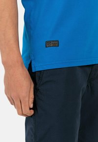 Niebieska koszulka z okrągłym dekoltem i krótkimi rękawami; wykonana z materiału bawełnianego; posiada małą czarną metkę na dolnym lewym boku. Noszona z ciemnymi szortami.