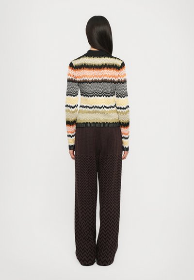 Maglione lavorato a maglia con motivi a zigzag in arancione, giallo, nero e crema; abbinato a pantaloni larghi a zigzag marroni; visto da dietro.