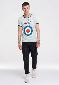 Light blue short-sleeve t-shirt featuring a graphic of Snoopy on a target design. Paired with black jeans and white sneakers, showcasing casual style.