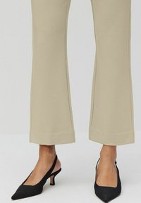 Beige utsvängda byxor med rak fåll, kombinerat med svarta spetsiga slingbackklackar, mot en neutral bakgrund.