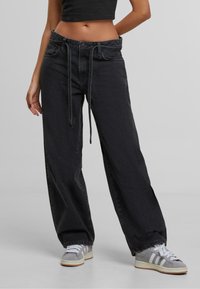 Henkilö, jolla on päällään löysät mustat farkut vyön kanssa, musta crop-top ja harmaa-valkoiset tennarit, seisoo harmaalla lattialla.