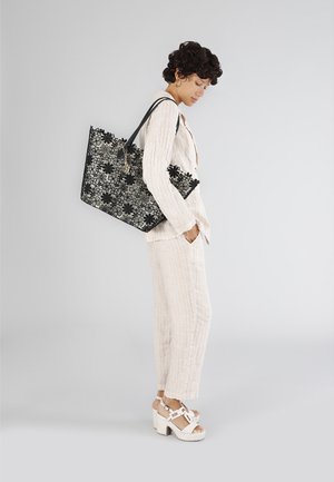 Zwarte bloemen tote bag met ingewikkeld kanten patroon, stevige handvatten en een platte bodem, vastgehouden door een persoon in een licht gestreepte outfit en witte schoenen.