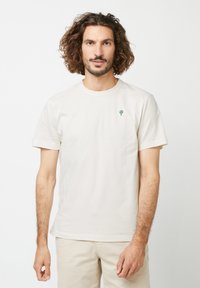 Helles beige Baumwoll-T-Shirt mit kurzen Ärmeln, Rundhalsausschnitt und einer kleinen grünen Baumstickerei auf der Brust, glatte Textur.