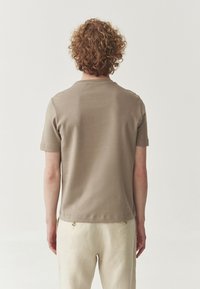 Tricou bej cu mânecă scurtă, cu decolteu rotund și textură netedă, văzut din spate, asortat cu pantaloni de o culoare deschisă.
