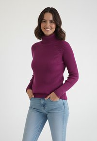 Fioletowy prążkowany sweter z golfem i długimi rękawami, o dopasowanym kroju. Nosi się go z jasnoniebieskimi dżinsami, ręce włożone w kieszenie.