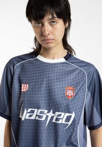 Zils sporta krekls ar režģa rakstu, kontrastējošo baltu apkakli, sarkaniem un baltiem logotipiem un īsām piedurknēm. Vieglas, elpojošas audumas.