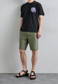 Homme portant un t-shirt graphique noir, un short vert olive et des sandales à plateforme noires se tenant devant un mur gris clair uni.
