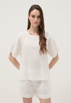 Jonge vrouw met lang bruin haar, gekleed in een losse witte korte mouwen blouse en witte shorts, staand met de handen achter haar rug.