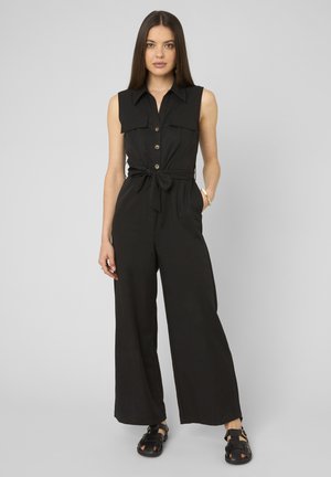 Femme aux longs cheveux bruns portant une combinaison noire sans manches avec des boutons, une ceinture à la taille, jambes larges et sandales noires, debout devant un fond gris.
