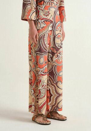 Persona che indossa pantaloni a gamba larga e un top a maniche lunghe abbinato con motivo paisley arancione, crema e marrone, abbinato a sandali marroni con perline.
