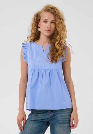 Mujer con cabello largo y rizado, vestida con una blusa azul sin mangas con volantes en los hombros y vaqueros azules sobre un fondo liso.
