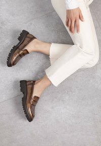 Mocasines de charol marrones con suela negra gruesa, que cuentan con una tira decorativa sobre el empeine, combinados con jeans claros desflecados.