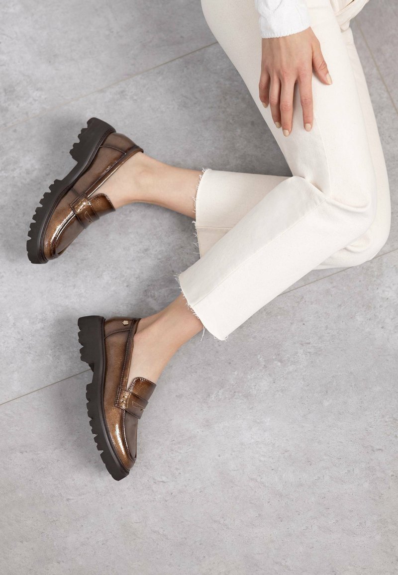 Mocasines de charol marrones con suela negra gruesa, que cuentan con una tira decorativa sobre el empeine, combinados con jeans claros desflecados.