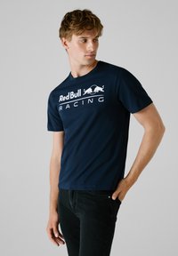 Navyblaues Baumwoll-T-Shirt mit dem weißen "Red Bull Racing"-Logo. Kurze Ärmel, Rundhalsausschnitt, glatte Textur und normale Passform.