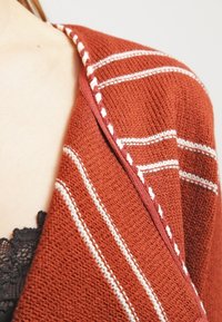 Kardigan z pleteného materiálu v barvě rezavé, s bílými pruhy, texturovanou látkou a kontrastním lemem podél okrajů. Otevřený střih, bez zapínání.