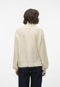 Krämfärgad stickad tröja med hög krage, avslappnad passform, sänkta axlar och ribbade muddar, i kombination med mörka jeans.