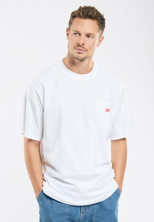 Man wearing a loose white T-shirt with a small red logo on the left chest pocket and blue jeans, hands in pockets, looking to the side.