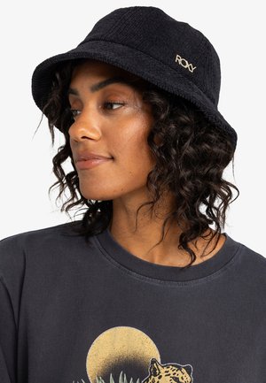 Woman with curly hair wearing a black textured bucket hat and dark graphic t-shirt featuring a leopard and yellow sun.