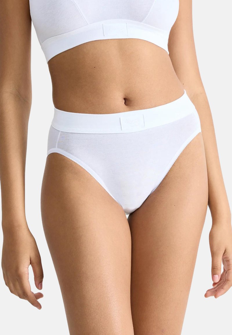 Frau trägt weiße High-Waist-Slips und einen passenden weißen Sport-BH, steht vor einem schlichten hellen Hintergrund.