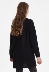 Černý pletený cardigan volného střihu s otevřeným vzorem, dlouhými rukávy a delším zadním lemem se bočními rozparky.