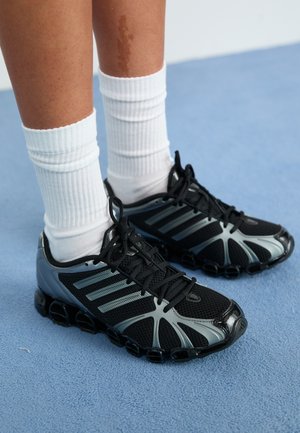 Persoană care poartă șosete albe cu dungi și adidași atletici negri cu accente gri pe podea cu mochetă albastră.