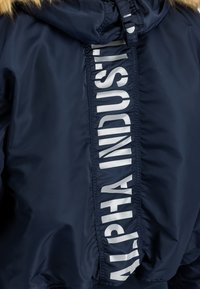 Marineblaue Jacke mit Kunstpelzbesatz an der Kapuze und einer vertikalen Reißverschlussabdeckung mit weißem "ALPHA INDUSTRIES" Schriftzug.