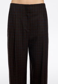 Pantaloni a quadri marroni con un motivo a griglia blu, pieghe e tasche laterali. Il tessuto ha una texture liscia e una vestibilità ampia.