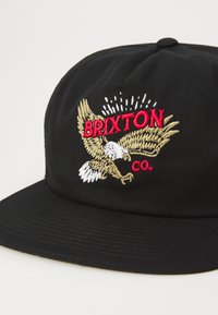 Sort kasket med en broderet design af en ørn og ordet "BRIXTON" i rødt. Stoffet har en tekstureret overflade med en struktureret form.