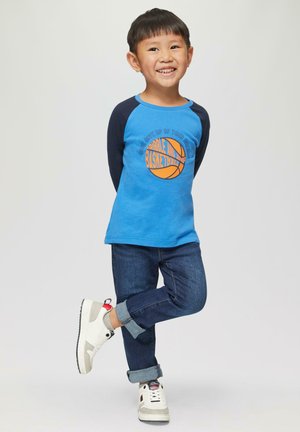 Blauw longsleeve t-shirt met een oranje basketbalafbeelding, gecombineerd met donker denim jeans en witte sneakers met grijze en rode accenten.