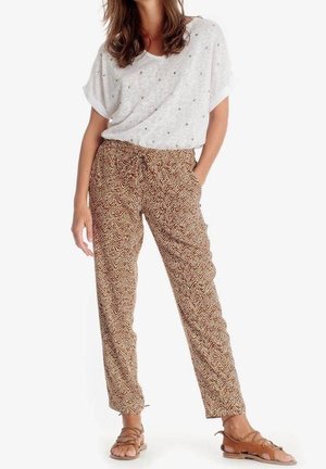 Vrouw die een wit shirt met korte mouwen en kleine stippen draagt, een bruine broek met een zigzagpatroon en trekkoorden in de taille, en bruine sandalen met bandjes.