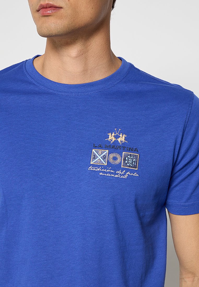 Blaues T-Shirt mit gesticktem La Martina-Logo, das zwei Polo-Spieler, drei dekorative Embleme und kursiven Text auf der linken Brust zeigt.