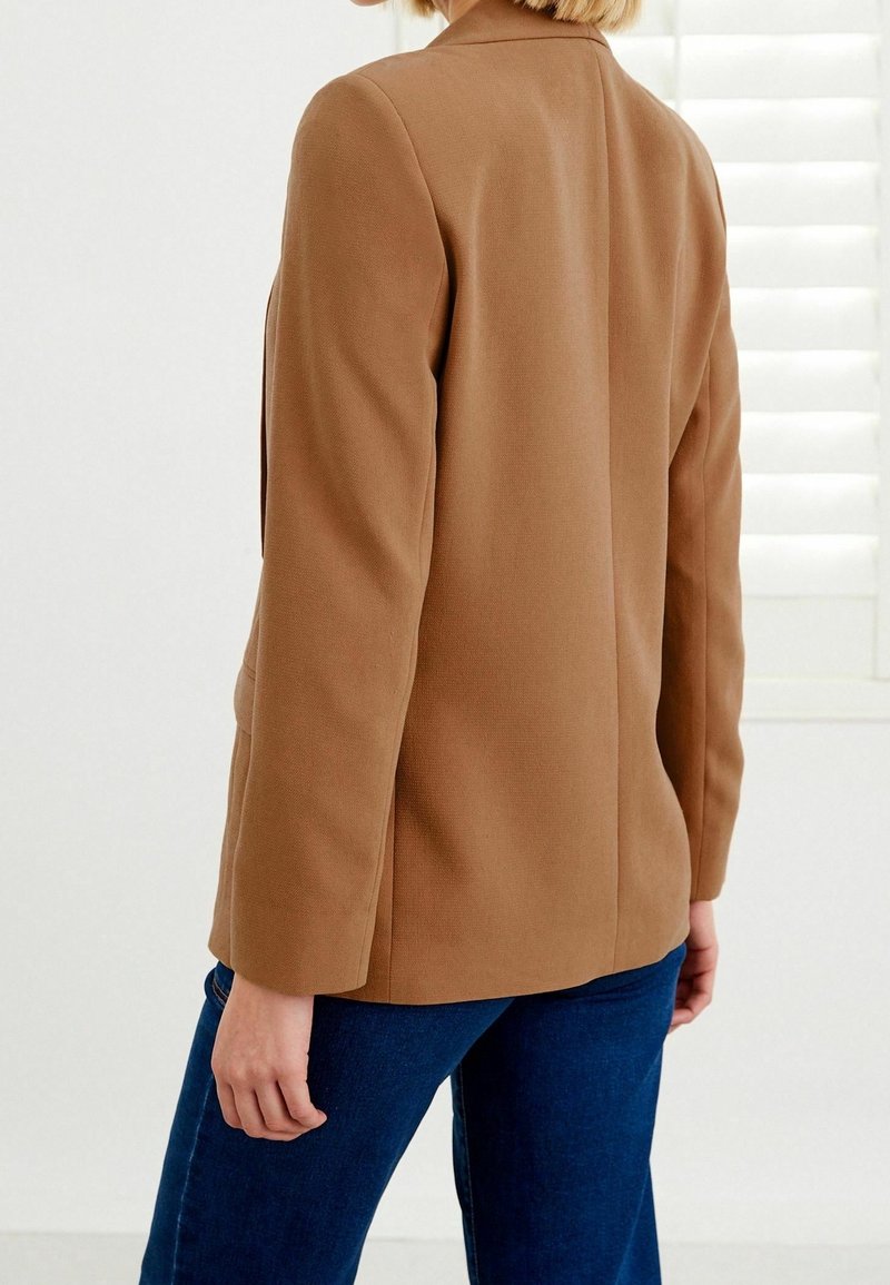 Personne portant un blazer camel et un jean bleu foncé, vue de dos contre un mur blanc avec des stores.