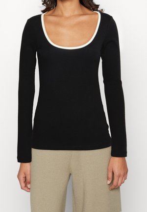 Femme portant un haut noir à manches longues avec un col échancré bordé de blanc et un pantalon beige, debout avec les bras détendus le long du corps.