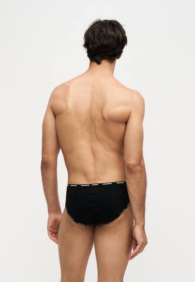 Homme torse nu vu de dos portant un slip noir avec une ceinture de marque sur fond clair uni.