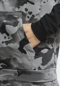 Sweat-shirt camouflage gris avec poche kangourou, tissu texturé et manche noire contrastante. Gros plan sur une main reposant dans la poche.