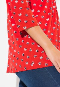 Rotes Baumwollhemd mit Dreiviertelärmeln, das mit einem Muster aus schwarzen und weißen Punkten versehen ist. Zeigt Nahtdetails an der Seite und eine glatte Textur.