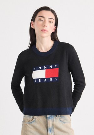 En ung kvinne med langt mørkt hår har på seg en svart genser med "TOMMY JEANS"-logoen. Hun står mot en lys bakgrunn.