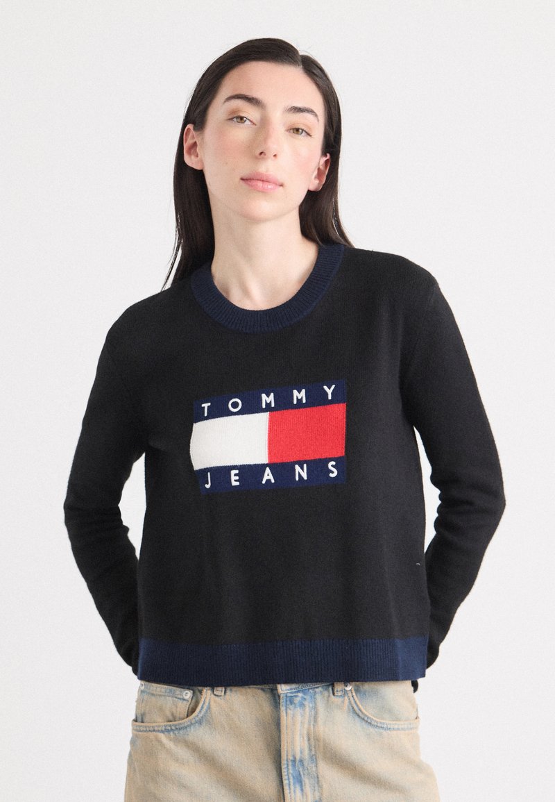 Une jeune femme aux longs cheveux noirs porte un pull noir arborant le logo "TOMMY JEANS". Elle se tient devant un fond clair.