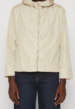 Veste à capuche beige clair avec zip, portée avec un jean bleu foncé, visible des épaules aux hanches sur un fond uni.