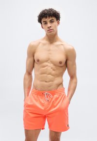 Νεαρός άνδρας με σγουρά μαλλιά που φοράει φωτεινά πορτοκαλί μαγιό, όρθιος με τα χέρια στις τσέπες μπροστά σε ένα απλό λευκό φόντο.