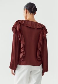 Brown long-sleeve blouse featuring a ruffled neckline and sleeves, made from lightweight fabric, with a relaxed fit and smooth texture.