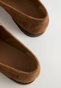Paire de mocassins sans lacets en daim marron avec semelles noires, vue de dessus sur une surface claire.