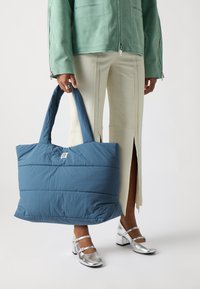 Sac fourre-tout rembourré bleu avec une texture lisse et une étiquette avec logo, doté de deux poignées robustes et de détails matelassés, porté avec un pantalon clair.