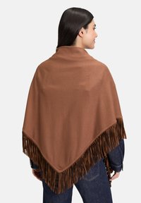 Femme aux longs cheveux foncés portant un châle à franges marron sur une veste en jean, de dos et souriante, sur un fond clair.