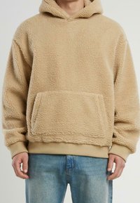 Sudadera de felpa beige con una superficie texturizada y suave, un bolsillo tipo canguro en la parte frontal y puños y dobladillo acanalados. Diseñada en un corte holgado.
