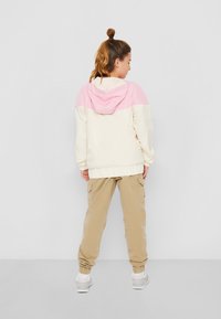 Lättvikts, krämfärgad jacka med huva, rosa övre del, elastiska ärmslut och midjeband, i kombination med beiga cargobyxor. Stickad textur.