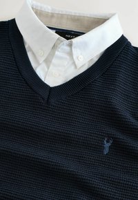 Marineblauwe gebreide pullover met een V-hals, gedragen over een witte geborduurde shirt. Bevat een textuurstof en een klein geborduurd hert-logo op de borst.