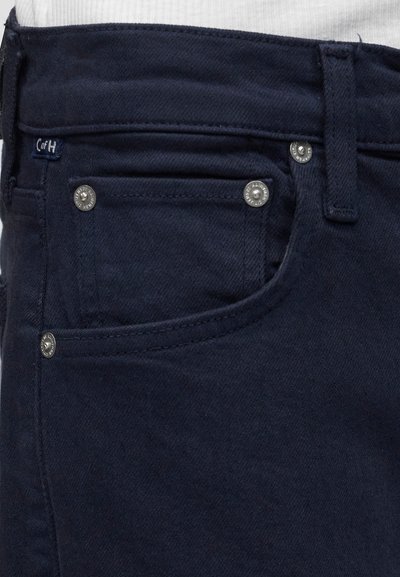 Κοντινή λήψη τσέπης από σκούρο μπλε τζιν με μεταλλικά τρουκς, θηλιά ζώνης και μικρή υφασμάτινη ετικέτα μάρκας.