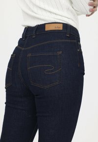 Dunkelblaue Denim-Jeans mit glatter Textur, versehen mit orangefarbenen Nähten, zwei Gesäßtaschen und einem hellbraunen Lederpatch am Bund.